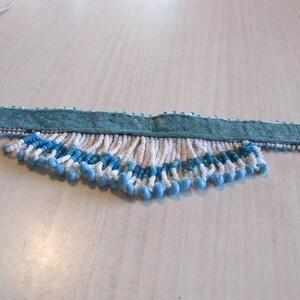 May include: A white and blue beaded choker necklace with a teal suede backing and white ribbon ties. The beads are arranged in a fringe pattern.