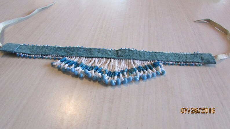 May include: A white and blue beaded choker necklace with a teal suede backing and white ribbon ties. The beads are arranged in a fringe pattern.