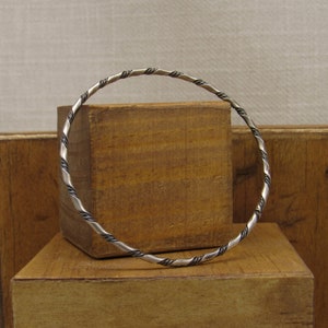 May include: A silver-toned bangle bracelet with a twisted rope design. The bracelet has alternating silver and dark gray sections, creating a textured pattern. The jewelry is displayed on a wooden block.