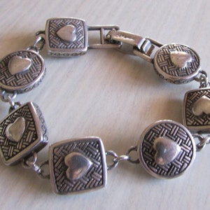 K&ouml;nnte beinhalten: Silberarmband mit einer Kette und mehreren herzf&ouml;rmigen Anh&auml;ngern. Jeder Anh&auml;nger hat ein gewebtes Muster und ein erhabenes Herz in der Mitte.