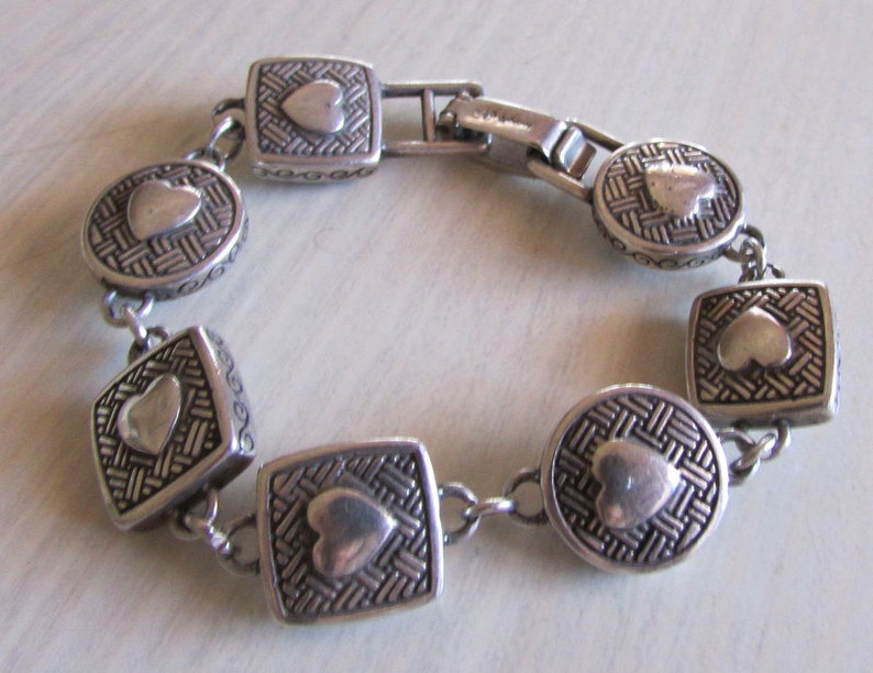 K&ouml;nnte beinhalten: Silberarmband mit einer Kette und mehreren herzf&ouml;rmigen Anh&auml;ngern. Jeder Anh&auml;nger hat ein gewebtes Muster und ein erhabenes Herz in der Mitte.