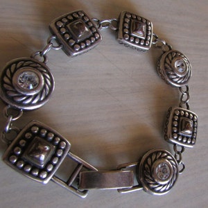 K&ouml;nnte beinhalten: Silberarmband mit einem Verschluss und dekorativen quadratischen und runden Gliedern mit klaren Strasssteinen.