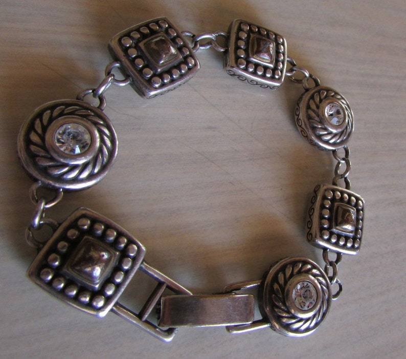 K&ouml;nnte beinhalten: Silberarmband mit einem Verschluss und dekorativen quadratischen und runden Gliedern mit klaren Strasssteinen.