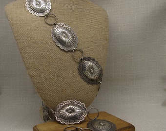 May include: A silver chain belt with five oval-shaped, embossed, silver medallions. The belt is displayed on a brown fabric-covered mannequin. The belt is also shown on a wooden surface with two additional medallions.
