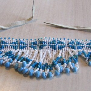 May include: A white and blue beaded choker necklace with a fringe of white and blue beads. The necklace is made of small, round beads and has a geometric pattern.