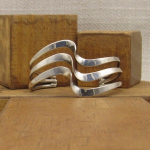 May include: A silver cuff bracelet with a unique, wavy design. The bracelet features four parallel, curved bands that create an open, flowing aesthetic. The polished metal reflects light, highlighting the bracelet's smooth, modern style. This piece is a statement accessory.