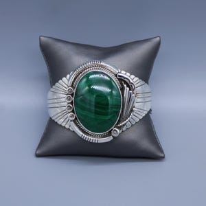 May include: A wide silver cuff bracelet featuring a large, oval-shaped green malachite stone at the center. The silver band is decorated with intricate stamped patterns, including sunburst motifs and twisted wire detailing around the stone.