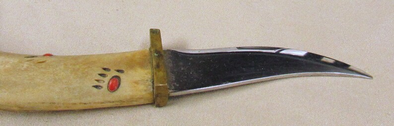 May include: A curved, black-handled knife with a bone handle. The handle is decorated with a red and black bear paw print.