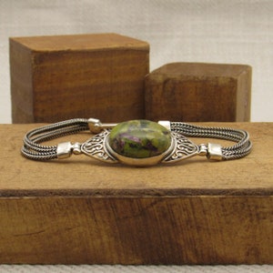 May include: A silver bracelet with a green and purple gemstone set in a silver filigree design. The bracelet has a braided chain design.