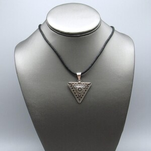 May include: A silver-colored triangular pendant necklace with a black cord. The pendant features an intricate geometric design with a central circular element and detailed edges. The necklace is displayed on a gray mannequin.