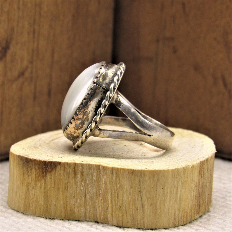 Puede incluir: Un anillo de plata con una piedra ovalada blanca engastada en un engaste de plata decorativo. El anillo tiene una banda ancha con un dise&ntilde;o retorcido.