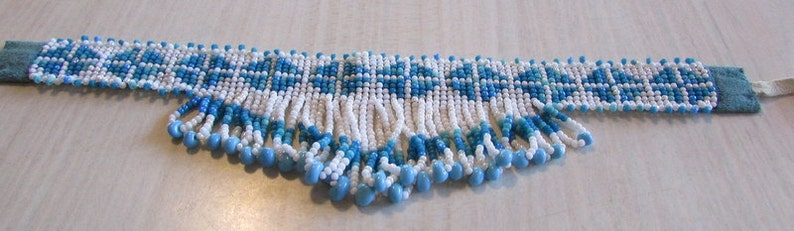 May include: A white and blue beaded necklace with a fringe of beads. The necklace is made of small, round beads and has a geometric pattern.