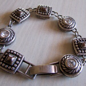 K&ouml;nnte beinhalten: Silberarmband mit einem Kettenglieddesign und quadratischen und runden Anh&auml;ngern mit klaren Strasssteinen. Die Anh&auml;nger haben ein strukturiertes, verziertes Design mit einem goldenen Akzent.