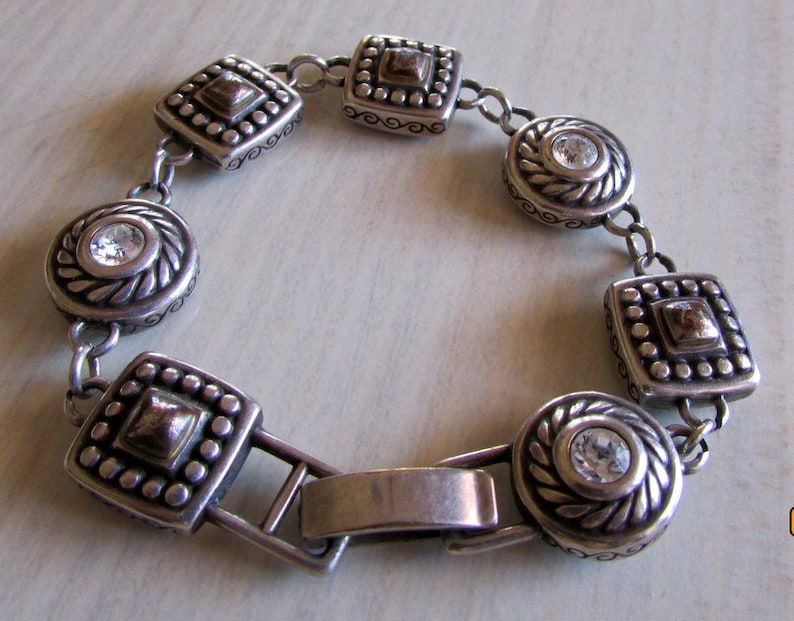 K&ouml;nnte beinhalten: Silberarmband mit einem Kettenglieddesign und quadratischen und runden Anh&auml;ngern mit klaren Strasssteinen. Die Anh&auml;nger haben ein strukturiertes, verziertes Design mit einem goldenen Akzent.