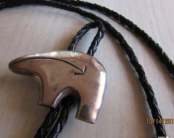 May include: A silver bolo tie featuring a stylized bear design. The tie is on a black braided leather cord.