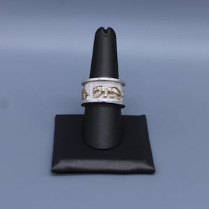 May include: A wide band ring featuring a textured silver finish and a series of raised, polished gold-toned panther figures encircling the center. The ring has smooth, polished silver edges at the top and bottom, creating a distinct contrast in color and texture.