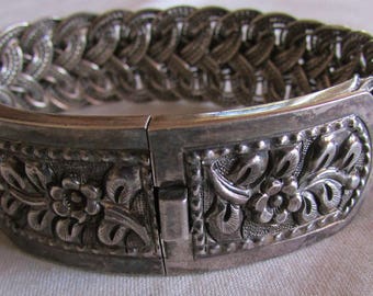 May include: A silver bracelet with a braided design and floral accents. The bracelet has a hinged clasp.