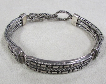 May include: A silver bracelet with a braided chain design and a decorative clasp. The bracelet has a textured, patterned band.