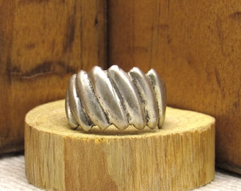 May include: A silver-colored ring with a ridged design, resting on a small wooden block. The ring's surface has a slightly aged appearance. The background features a wooden surface, providing a rustic contrast to the jewelry.