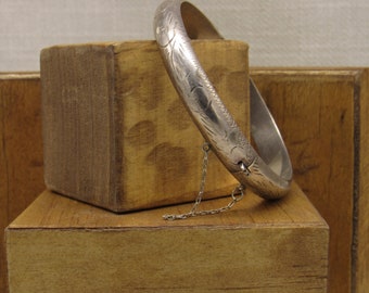 May include: A silver bangle bracelet with an engraved floral design. The bracelet has a hinged clasp and a delicate chain attached.
