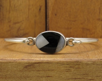 May include: A silver bangle bracelet with a black and white oval inlay. The inlay is made of mother of pearl and onyx.