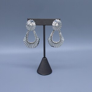 May include: A pair of silver-toned earrings featuring a circular top with a sunburst pattern and a dangling teardrop-shaped base with a fan-like design. The earrings are displayed on a black T-shaped stand, showcasing a polished metallic finish.