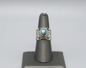 May include: A silver ring with a round, blue gemstone at the center. The ring has an ornate, openwork design around the gemstone. The ring is displayed on a black ring holder against a gray background.