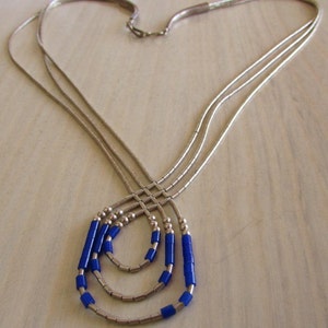 May include: A silver necklace with three strands, each adorned with small, bright blue beads. The necklace has a delicate design, with the strands converging at the clasp and the blue beads adding a pop of color.