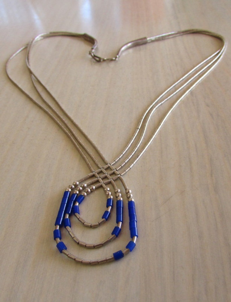 May include: A silver necklace with three strands, each adorned with small, bright blue beads. The necklace has a delicate design, with the strands converging at the clasp and the blue beads adding a pop of color.