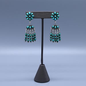 May include: Silver-toned dangle earrings with turquoise-colored stones. Each earring features a flower-shaped top and a fringe of turquoise beads. The earrings are displayed on a black jewelry stand against a light blue background.