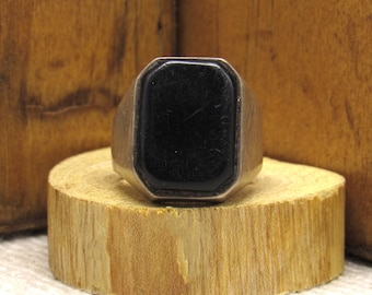 May include: A silver ring with a black rectangular stone set in the center. The ring is sitting on a wooden surface.