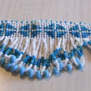 May include: A white and blue beaded necklace with a fringe of beads. The necklace has a geometric pattern.