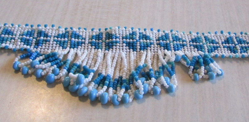 May include: A white and blue beaded necklace with a fringe of beads. The necklace has a geometric pattern.
