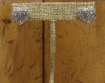 May include: A pair of silver heart-shaped earrings with a textured design. The earrings are displayed on a wooden stand with a burlap backing.
