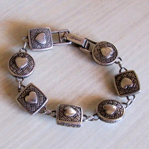 K&ouml;nnte beinhalten: Silberarmband mit einem Kettenglieddesign und herzf&ouml;rmigen Anh&auml;ngern. Jeder Anh&auml;nger hat ein gewebtes Muster und ein erhabenes Herz in der Mitte. Das Armband hat einen Verschluss mit der Aufschrift "Brighton".