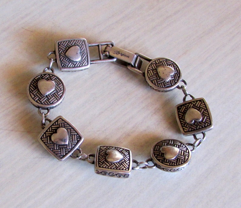 K&ouml;nnte beinhalten: Silberarmband mit einem Kettenglieddesign und herzf&ouml;rmigen Anh&auml;ngern. Jeder Anh&auml;nger hat ein gewebtes Muster und ein erhabenes Herz in der Mitte. Das Armband hat einen Verschluss mit der Aufschrift "Brighton".