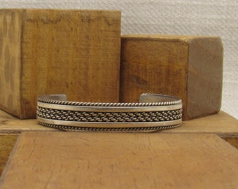 May include: A silver bracelet with a braided design. The bracelet has a rope-like edge and a textured band.