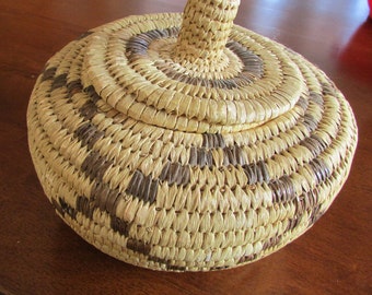May include: A round, woven basket with a lid. The basket is made of natural materials and has a brown and tan pattern. The lid has a raised center.