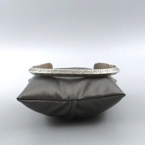 May include: A silver cuff bracelet with a geometric pattern. The bracelet is displayed on a gray pillow against a light gray background. The cuff is open and has a series of repeating triangular shapes etched around the band.