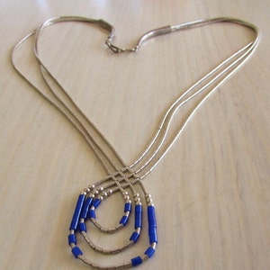 May include: A silver-toned necklace with three strands, each adorned with blue cylindrical beads. The necklace is arranged in a heart shape, with the strands converging at the clasp. The beads are a vibrant blue color.
