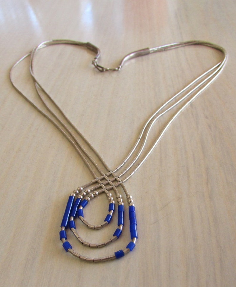 May include: A silver-toned necklace with three strands, each adorned with blue cylindrical beads. The necklace is arranged in a heart shape, with the strands converging at the clasp. The beads are a vibrant blue color.