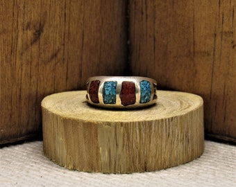May include: A silver ring with red and blue inlay stones. The ring is set on a wooden surface.