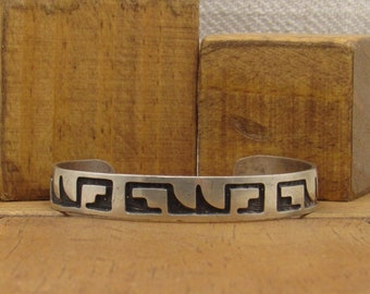 May include: A silver bracelet with a black geometric design. The bracelet is on a wooden surface.