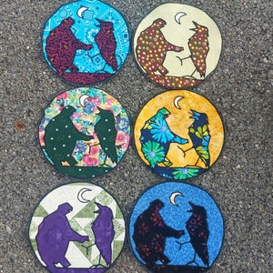 May include: Six round fabric coasters with a blue, yellow, or white background. Each coaster features a pair of black silhouettes of birds with a crescent moon in the upper right corner.