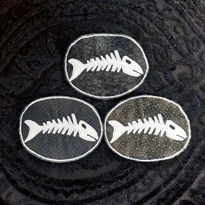 May include: Three black and white fish bone patches with a distressed look. The patches are oval shaped and have a white fish bone design on a black background.