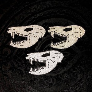 May include: Three white embroidered patches of animal skulls with black outlines. The skulls are facing forward with their mouths open.