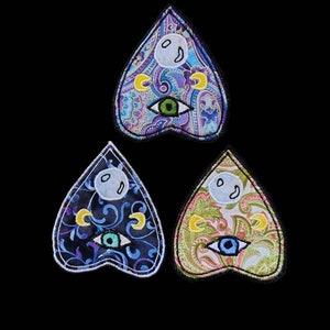 May include: Three heart-shaped patches with a paisley design. Each patch features a white ghost, a crescent moon, and a single eye. The patches are in shades of blue, green, and pink.