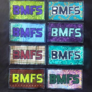 Puede incluir: Ocho parches de tela con las letras "BMFS" en diferentes colores y patrones. Los parches están cosidos sobre un fondo de tela negro.
