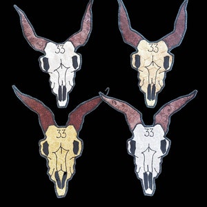 Puede incluir: Cuatro diseños decorativos de calaveras con cuernos marrones y el número "33" en la frente. Cada calavera tiene una combinación de colores diferente: blanco, beige, dorado y plateado. El fondo es negro.