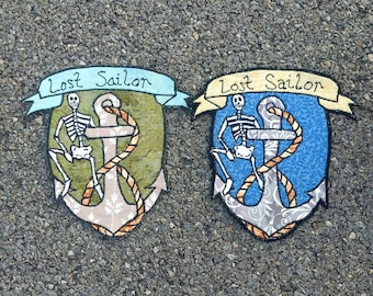 May include: Two embroidered patches with a skeleton holding an anchor. The patches are on a gray gravel background. The text on the patches reads "Lost Sailor".